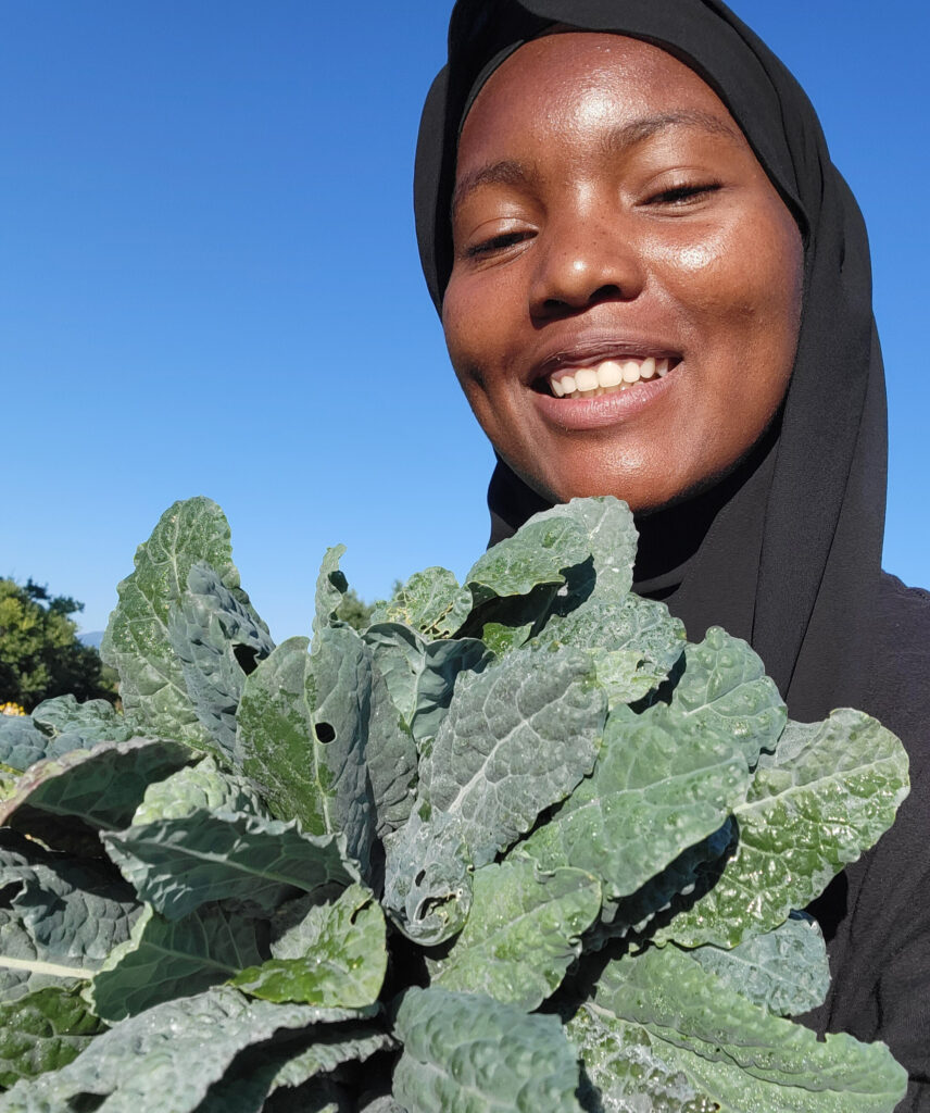 Headshot of Saleema wearing a black hijab and holding a large bushel of kale.